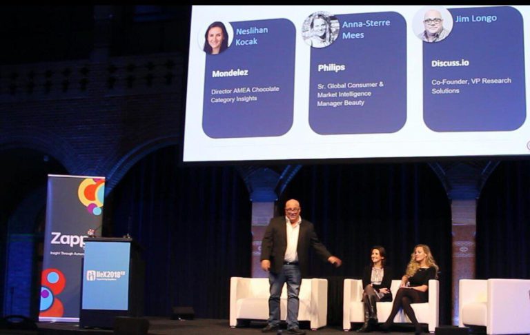 consumer centricity A speaker stands in front of a projected presentation slide with three seated panelists on stage during a conference.