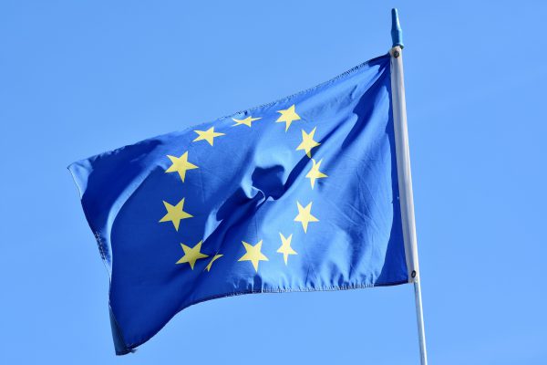 European Union flag with yellow stars on a blue background, waving on a flagpole against a clear blue sky.