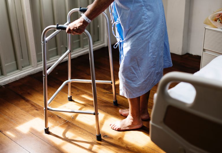 logo_IIEXHealth A person in a hospital gown uses a walker beside a bed on wooden flooring.
