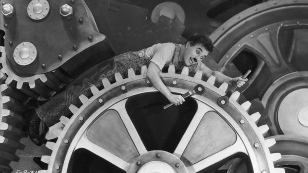 A person in overalls is caught in large industrial gears, holding wrenches, with an expression of surprise.