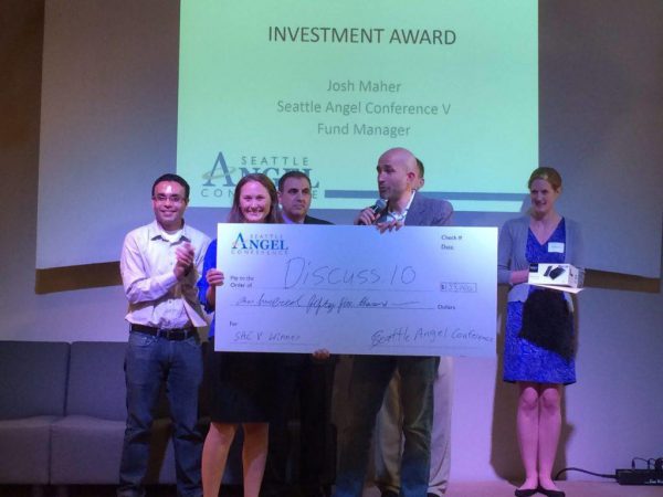 Group of people standing on stage, one holding a large check for Discuss.IO as part of the Seattle Angel Conference Investment Award. A presentation screen is in the background.