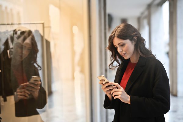 Woman in a black coat looks at her smartphone while standing near a glass window, her reflection visible.