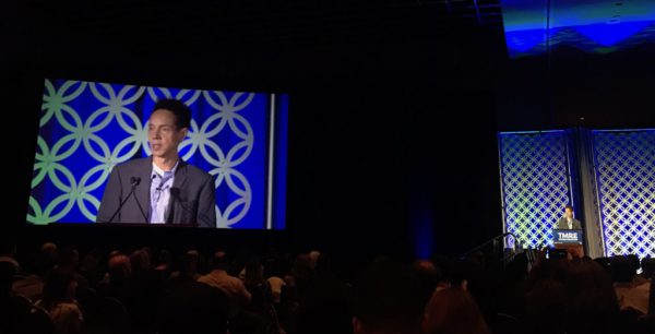 A speaker addresses an audience from a stage with a blue geometric backdrop. A large screen displays his image to the left.