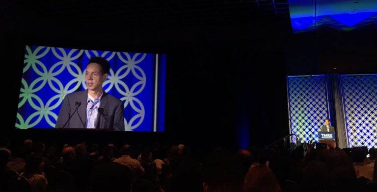 gladwell-tmre A speaker addresses an audience from a stage with a blue geometric backdrop. A large screen displays his image to the left.