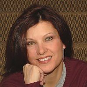 karen-1 Person with shoulder-length dark hair and earrings, smiling, rests their chin on their hand against a patterned staircase background.