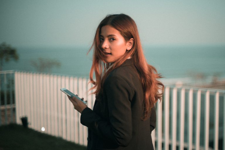 Consumer Centricity A woman with long hair wearing a dark jacket stands outdoors, holding a smartphone, with a fence and hazy water view in the background.