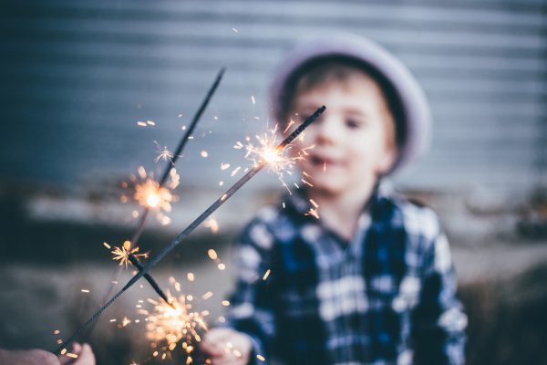Child in a plaid shirt and hat holding sparklers, with a blurred background.