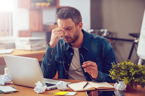 Man sits at a desk, holding his forehead and glasses, looking stressed. A laptop and crumpled papers are on the desk.