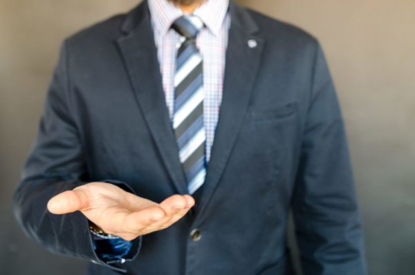 A person in a suit extends their open hand against a blurred, neutral background.