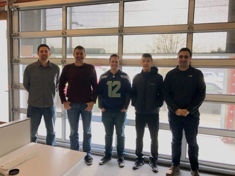 new hires Five people standing in a row indoors, smiling at the camera. They are in front of a large garage door with windows.