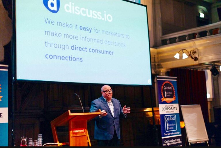 Consumer Closeness A speaker presenting at a podium in front of a screen displaying a slide about marketing with Discuss.io in a lecture hall.