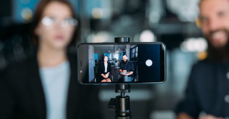 Video interview A smartphone on a tripod captures two people sitting and discussing, one holding a tablet to review the voice of the customer. The background is artistically blurred.