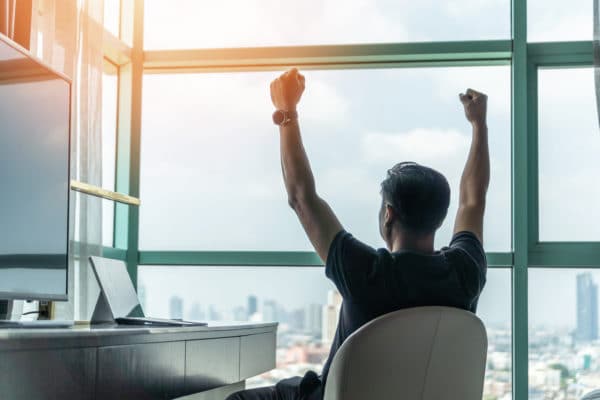 A person sits at a desk facing a large window with arms raised, overlooking a cityscape.