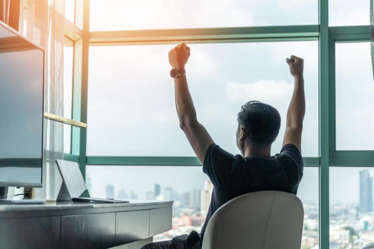 Business,Achievement,Concept,With,Happy,Businessman,Relaxing,In,Office,Or A person sits at a desk facing a large window with arms raised, overlooking a cityscape.