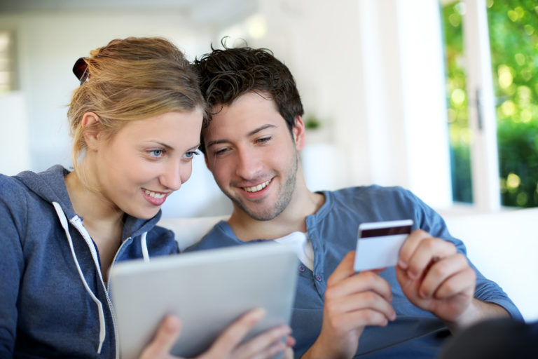 Happy,Young,Couple,Doing,Shopping,On,Internet A couple sits on a couch, smiling and looking at a tablet. The man holds a credit card. Bright room with a window in the background.