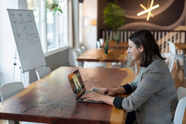 Woman seated at a wooden table, using a laptop for a video call in a well-lit room with whiteboard and decor.