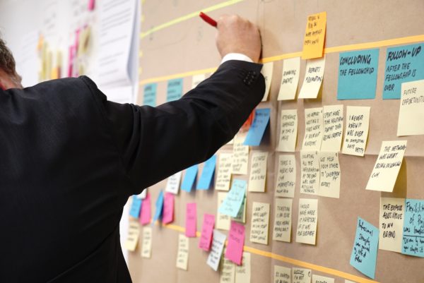 Person in a suit writes on a corkboard covered with multicolored sticky notes and lined paper.