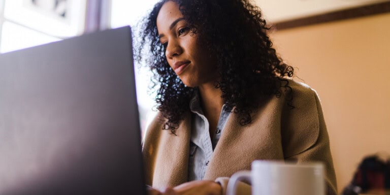 A person with curly hair focuses intently on their laptop, conducting AI-moderated research, with a steaming mug sitting prominently in the foreground on the table.