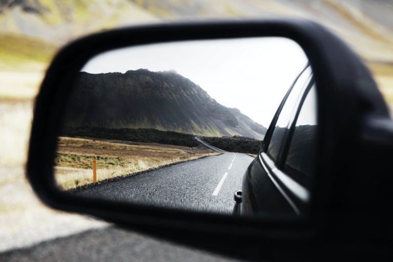 rearview Rearview mirror reflecting a long, empty road stretching into the distance, with mountains visible under a cloudy sky.