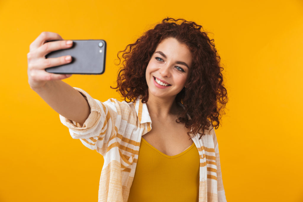 A woman with curly hair smiles while taking a selfie against a bright yellow background. She is wearing a striped shirt and a yellow top.