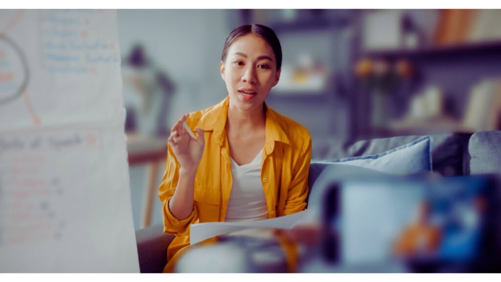 A woman in a yellow shirt sits on a couch, talking to a camera. A blurred chart is visible in the foreground.