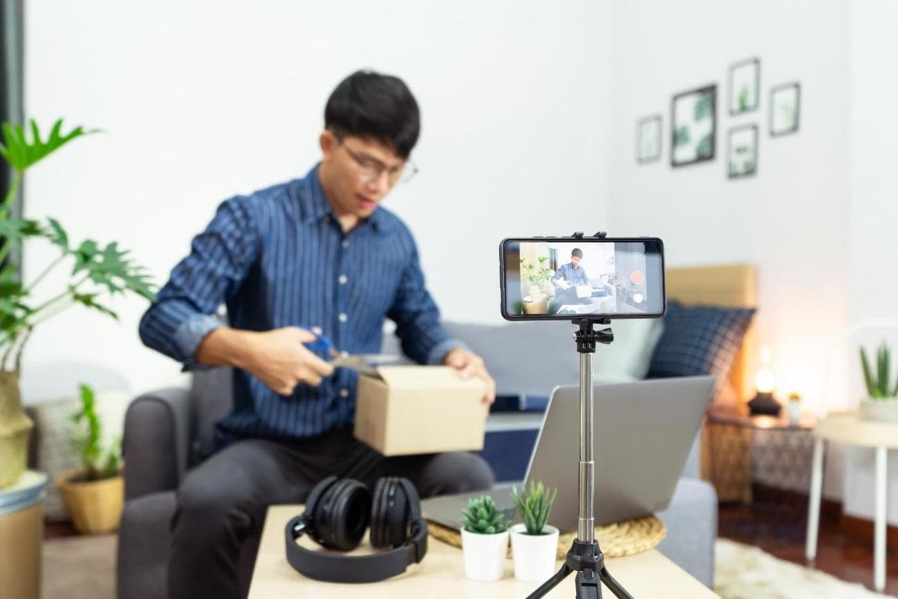 Unboxing Person in striped shirt opens a box with scissors while being recorded by a smartphone on a tripod. Headphones, plants, and a laptop are on the table nearby.