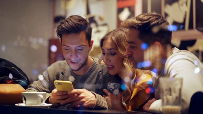 Three people sit closely at a cafe table, diving into consumer insights research as they examine data on a smartphone together.