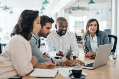 Four people in a meeting room are looking at a laptop screen, with notebooks and cups on the table.