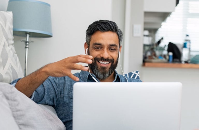 shutterstock_1297116592 A man with headphones smiles while gesturing at a laptop screen in a home setting.