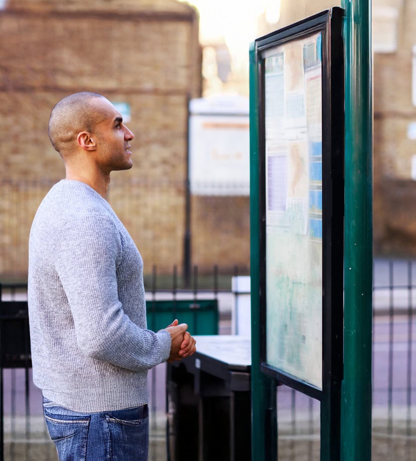 Group 2019912085664465 1 A man in a gray sweater and jeans reads a bulletin board outdoors.
