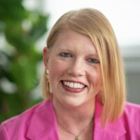 Person with straight blonde hair wearing a bright pink blazer smiles at the camera. There is a green plant blurred in the background.
