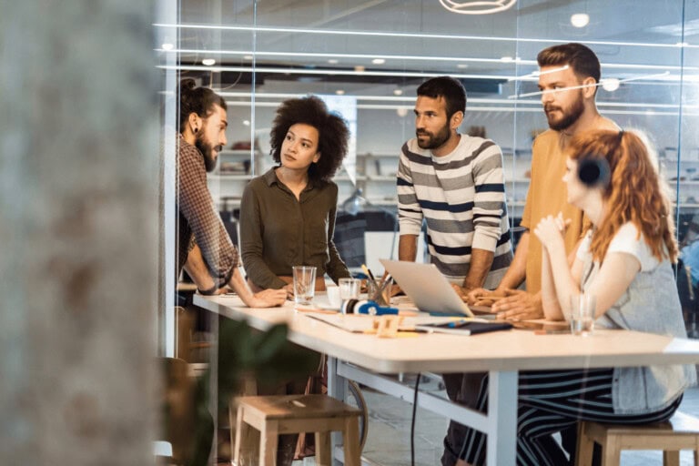 A group of people having a discussion around a table in a modern office setting, with laptops and notebooks.