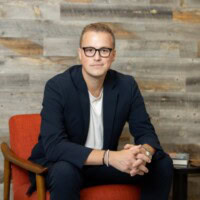 A person wearing glasses and a dark blazer sits on an orange chair. A wooden wall is in the background.