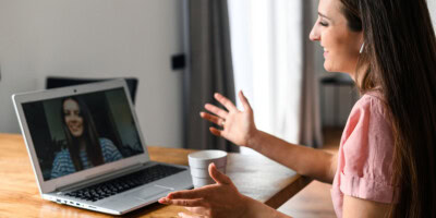 A woman sits at a table engaged in a video call on a laptop, with a cup of coffee nearby.
