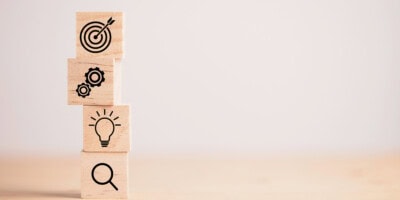 Four wooden blocks stacked vertically display icons of a target, gears, a light bulb, and a magnifying glass from top to bottom.