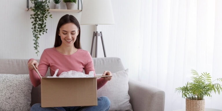 Blog headers A woman sits on a sofa, smiling as she opens a cardboard box.