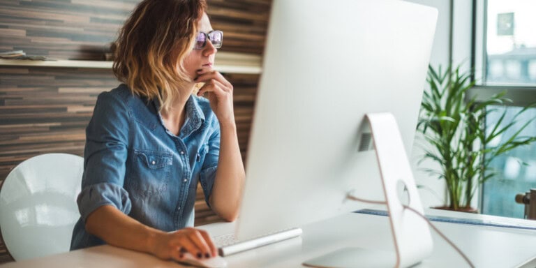 Qual data analysis Person wearing glasses and a denim shirt is sitting at a desk, thoughtfully looking at a computer screen.