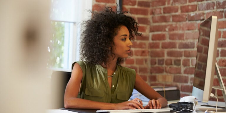 Techniques A woman with curly hair works at a computer in an office with brick walls. She is wearing a green sleeveless top.