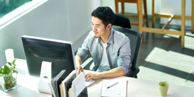Man working on a computer at a desk with books, a notebook, and a green mug, near a bright window.