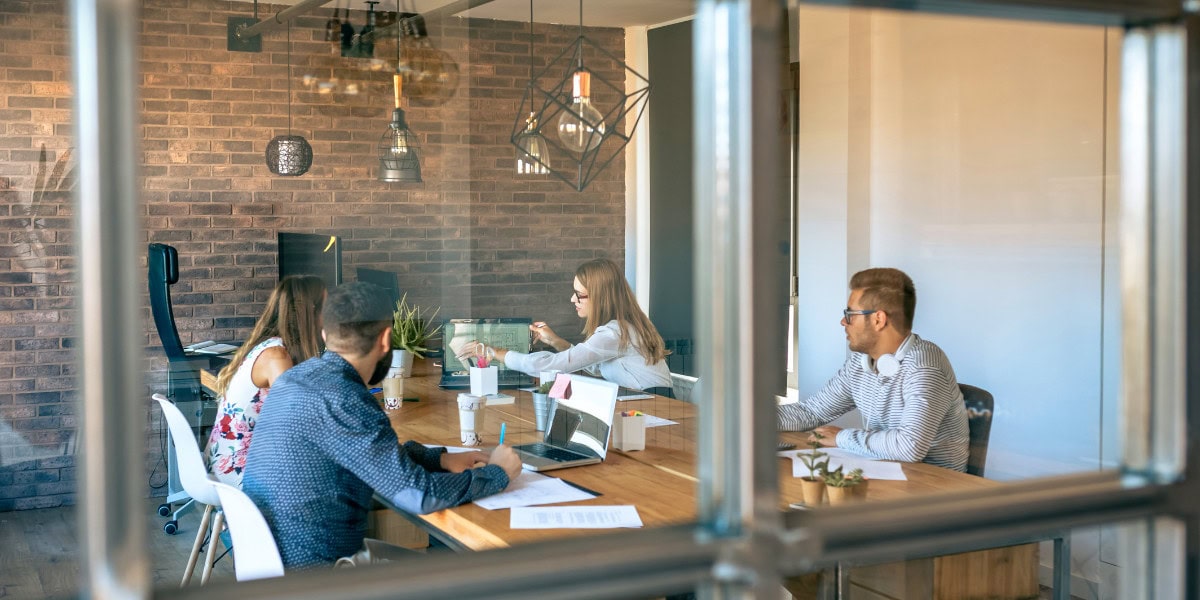 Team collaboration Four people engaged in a meeting around a wooden table in a modern office with brick walls, as seen through a glass partition.