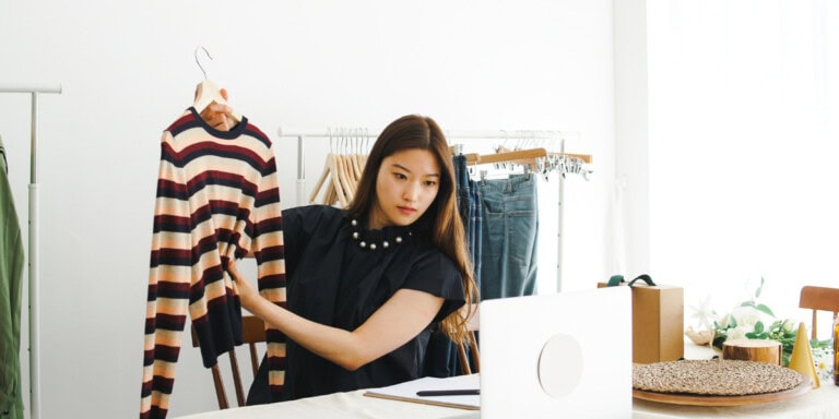 AI Moderated Feedback Systems- A Game-Changer for Business Growth A woman holds up a striped sweater in front of a laptop, surrounded by clothes on racks and a table with a circular woven mat.