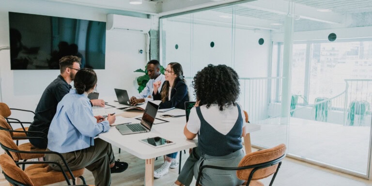 Benefits of AI Moderated Feedback for Real-Time Decision-Making Five people in a modern conference room engaged in discussion around a table with laptops and notes.