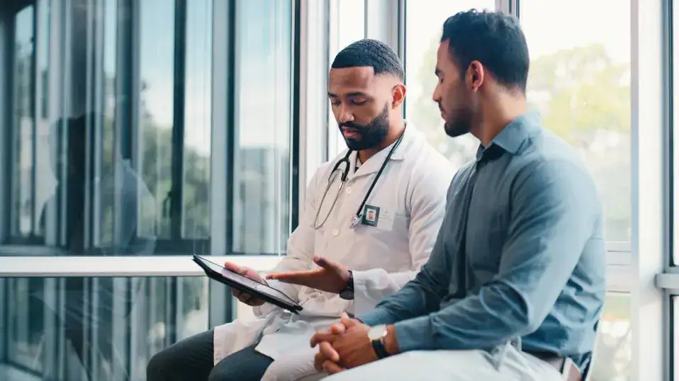A doctor in a white coat shows a tablet to a man in a blue shirt as they sit and discuss something in a modern office setting.