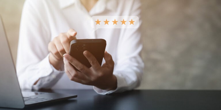 Blog headers A person in a white shirt uses a smartphone, with a five-star rating graphic above the device; a laptop is on the table nearby.