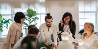 Five people gather around a table in a bright office, discussing work. There are plants and desk lamps in the room.