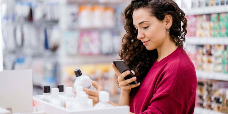 product A woman in a store examines a bottle of product while holding her smartphone, standing in an aisle with shelves of various items.
