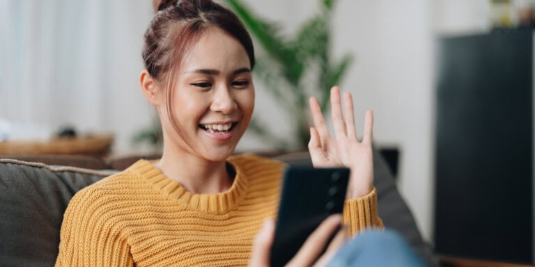 AI research tools A woman in a yellow sweater sits on a sofa, smiling and waving at her smartphone during a video call.