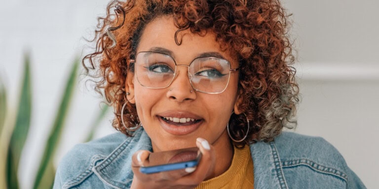 Blog headers A person with curly hair and glasses holds a smartphone and speaks into it, appearing engaged in conversation.