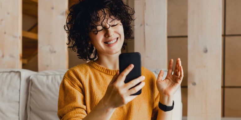 Blog3 A person with curly hair wearing a yellow sweater sits on a couch, smiling and waving at a smartphone during a video call.