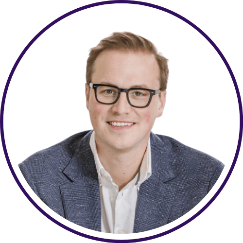 A man with short light hair and glasses is smiling, wearing a white shirt and a blue textured blazer, ready to share insights on Mastering GenAI, against a plain white background.